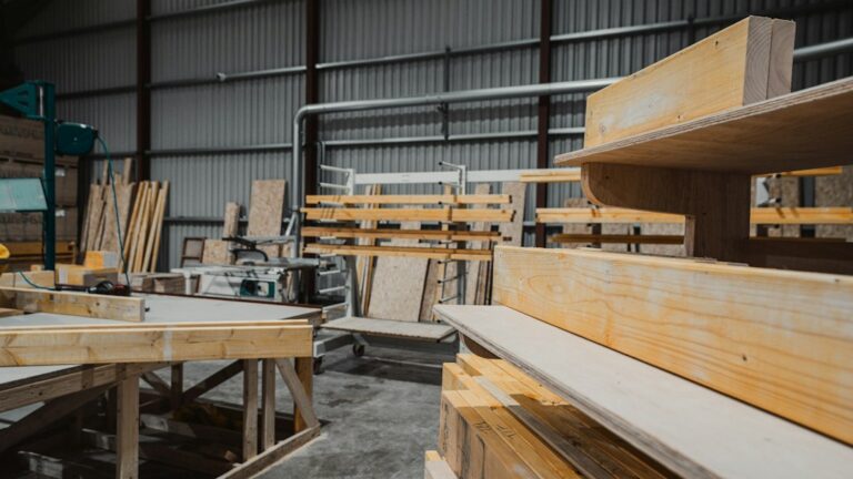 Straight and warped lumber boards arranged beside plywood panels and woodworking tools in a covered workspace.